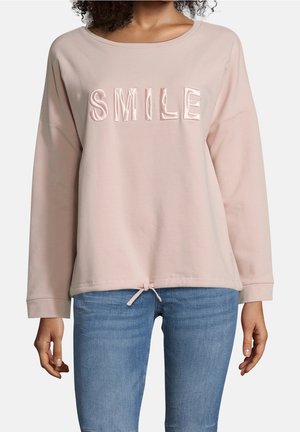 Vrouw draagt een lichtroze top met lange mouwen met ‘SMILE’ geborduurd op de borst en blauwe jeans, staat met ontspannen armen langs het lichaam.