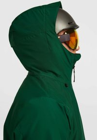 Groene waterdichte jas met een grote capuchon, zijrits en verstelbaar trekkoord. Gedragen met een zwarte helm en oranje getinte goggles.