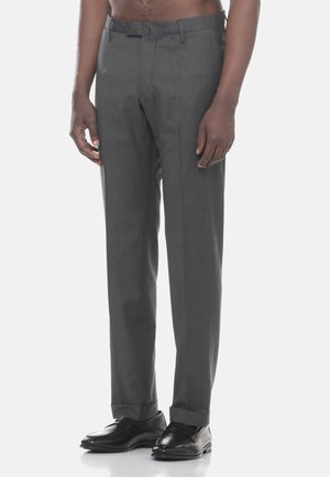 Uomo che indossa pantaloni eleganti grigi e mocassini neri in pelle, in piedi con le mani rilassate lungo i fianchi su uno sfondo chiaro e semplice.