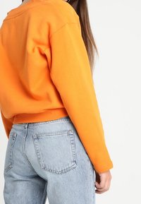 Femme portant un sweat-shirt orange vif et un jean bleu clair, montrée de dos et de la taille au milieu du torse sur fond blanc.