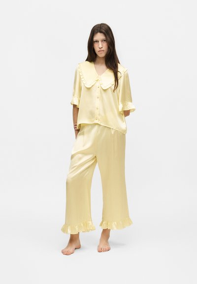 Femme debout pieds nus, portant un pyjama ample en satin jaune pâle avec un grand col à volants et des poignets à volants aux manches et aux ourlets du pantalon.