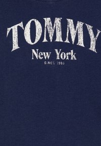 Marineblå T-skjorte laget av bomull, med hvit teksturert trykt tekst som leser "TOMMY New York" og "SINCE 1985" under.