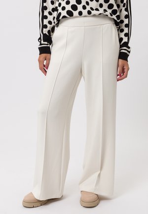 Pantalon à jambes larges en tissu crème avec une texture lisse. Dispose de plis à l'avant et d'une ceinture élastique, associé à des chaussures beige clair.