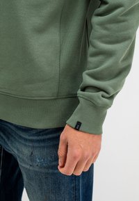 Groene pullover sweatshirt met geribde manchetten en kraag. Bevat een klein merklabel op de mouw. Draag met blauwe jeans, die lichte slijtage vertonen.