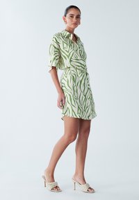 Kortærmet skjortekjole lavet af let stof med hvid baggrund og grønne abstrakte striber; har krave og knapdetaljer i siden.