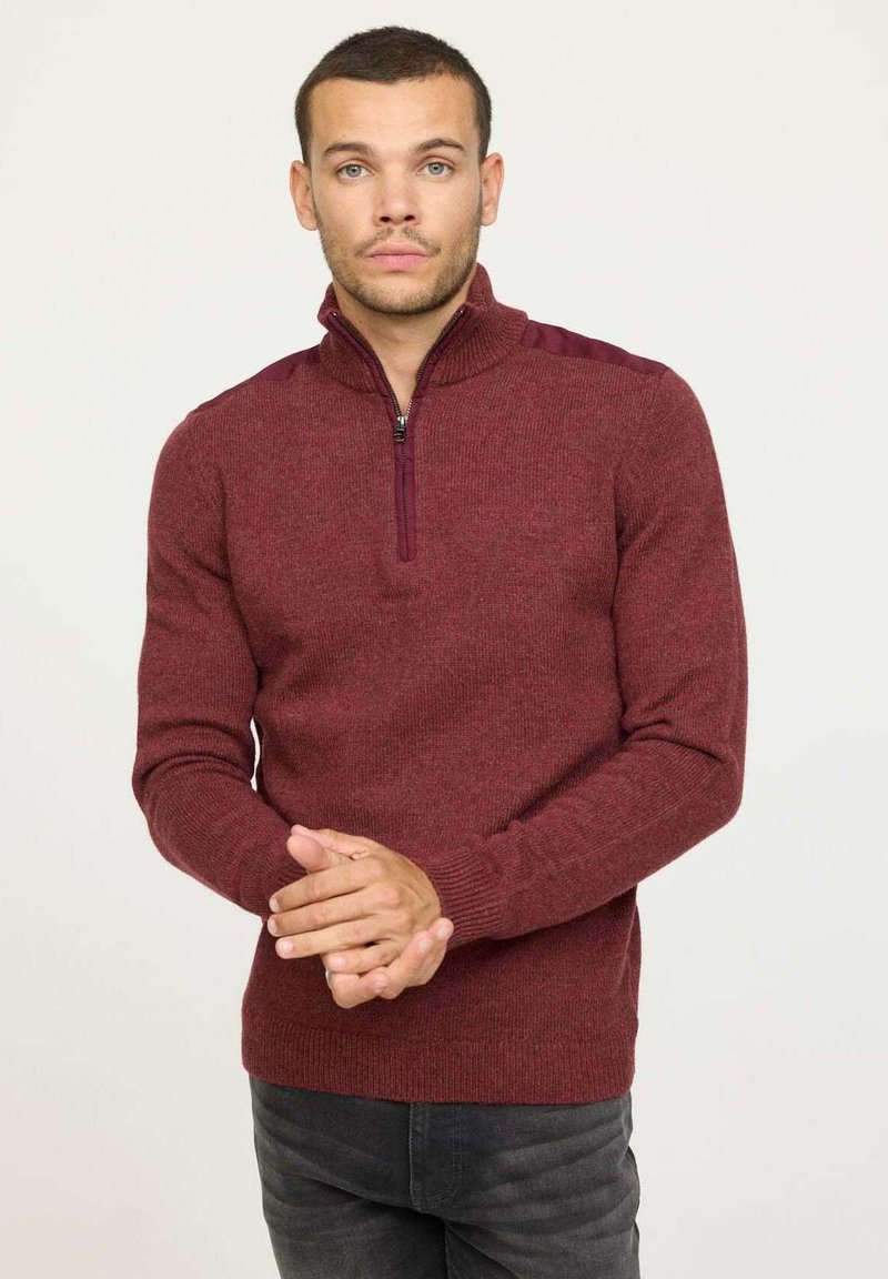 Pull-over bordeaux à fermeture éclair avec un tricot texturé, un col montant, et des accents contrastants sur les épaules, accompagné d'un jean foncé.