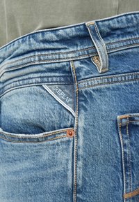 Μπλε τζιν με ξεθωριασμένη υφή, σχέδιο με πέντε τσέπες και ορατή ετικέτα "REPLAY" στην δεξιά πίσω τσέπη.