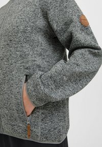 Graue Fleecejacke mit strukturiertem Muster, Reißverschlusstaschen und einem runden Lederpatch am Oberarm.