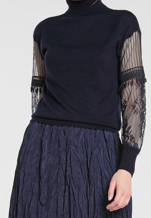 Femme portant un pull à col roulé bleu marine avec des manches en dentelle transparente et une jupe texturée bleu marine, debout avec une main détendue.