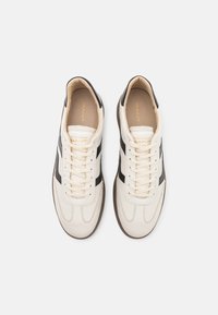 Hvite lær sneakers med svarte striper, rund tupp, flate lisser og brun gummisåle. Innvendig er de beige med gull merkevarelogo.