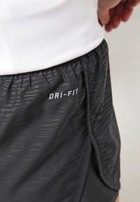 Gros plan sur un short de sport noir Dri-FIT avec un motif subtil, porté par une personne en chemise blanche, montrant la ceinture et la fente latérale.