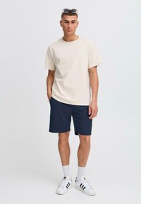 T-shirt en coton beige, shorts bleu marine, baskets blanches avec des rayures noires, et des lunettes de soleil sur la tête. Tenue simple et décontractée avec une coupe ample.