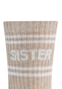 Kniestrümpfe in Beige mit weißen Streifen und dem Wort "SISTER" in Weiß seitlich eingestrickt. Hergestellt aus einem weichen, strukturierten Stoff.