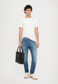 Λευκό βαμβακερό μπλουζάκι με λογότυπο, μπλε τζιν παντελόνι, κρέμα αθλητικά παπούτσια και μαύρη τσάντα tote με φερμουάρ, στραμμένο μπροστά σε ένα απλό φόντο.