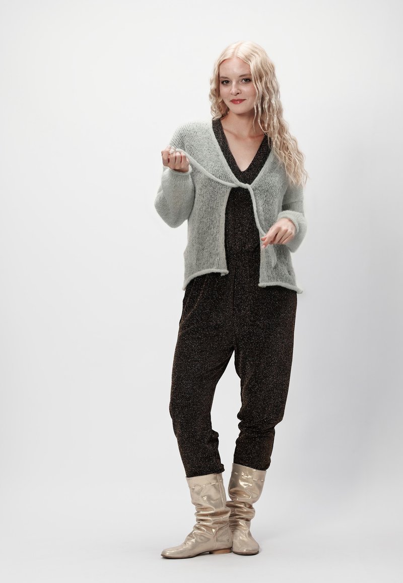 Jasnozielony, puszysty kardigan z zapięciem na wiązanie, noszony na czarnym kombinezonie z delikatnym blaskiem. Szare, luźne botki dopełniają całość.