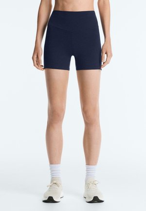 Vrouw die marineblauwe, high-waist sportshorts draagt, met witte enkelsokken en lichtbeige hardloopschoenen, staand op een witte achtergrond.