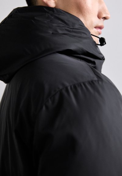 Jachetă pufă neagră cu guler înalt, material texturat și șnur ajustabil la glugă. Se prezintă cu un design elegant și modern.