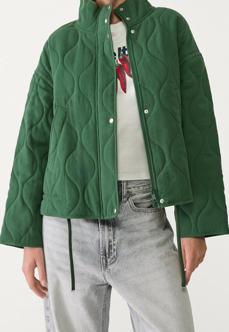 Veste matelassée verte à col montant portée ouverte sur chemise blanche avec imprimé de piments rouges, associée à un jean taille haute gris clair.