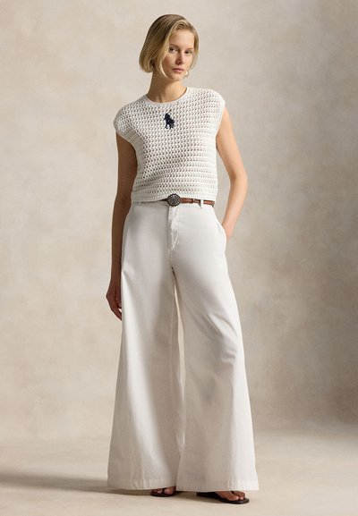 Γυναίκα που στέκεται με το ένα χέρι στην τσέπη φορώντας λευκό αμάνικο τοπ πλεκτό με σκουρόχρωμο λογότυπο, παντελόνι σε φαρδιά γραμμή λευκό, καφέ ζώνη και μαύρα σανδάλια.