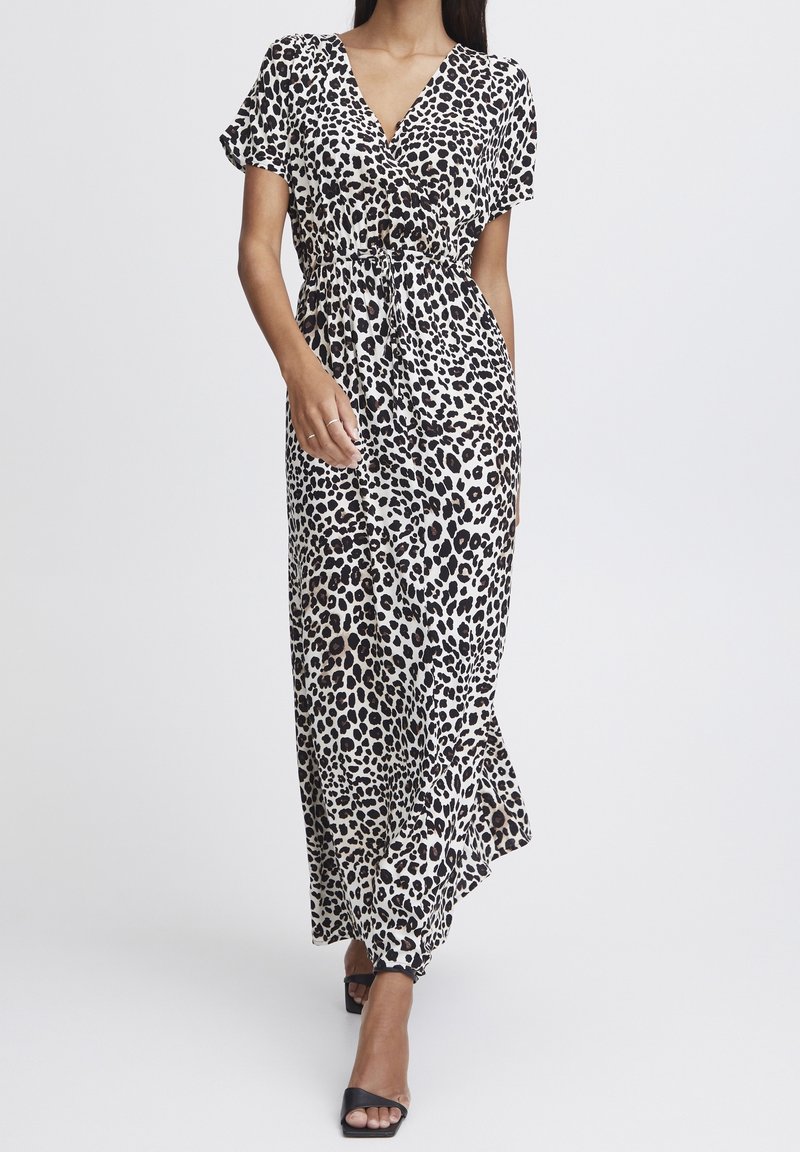 Femme portant une robe blanche longue jusqu'aux chevilles avec des taches léopard noires et brunes, un décolleté en V, des manches courtes, et des talons ouverts noirs.
