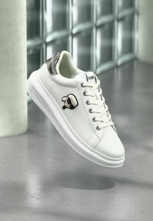 Hvid Karl Lagerfeld sneaker med snørebånd, sort og hvid Karl-logo på siden og metallisk hældetalje, vist mod en glasblokvæg.