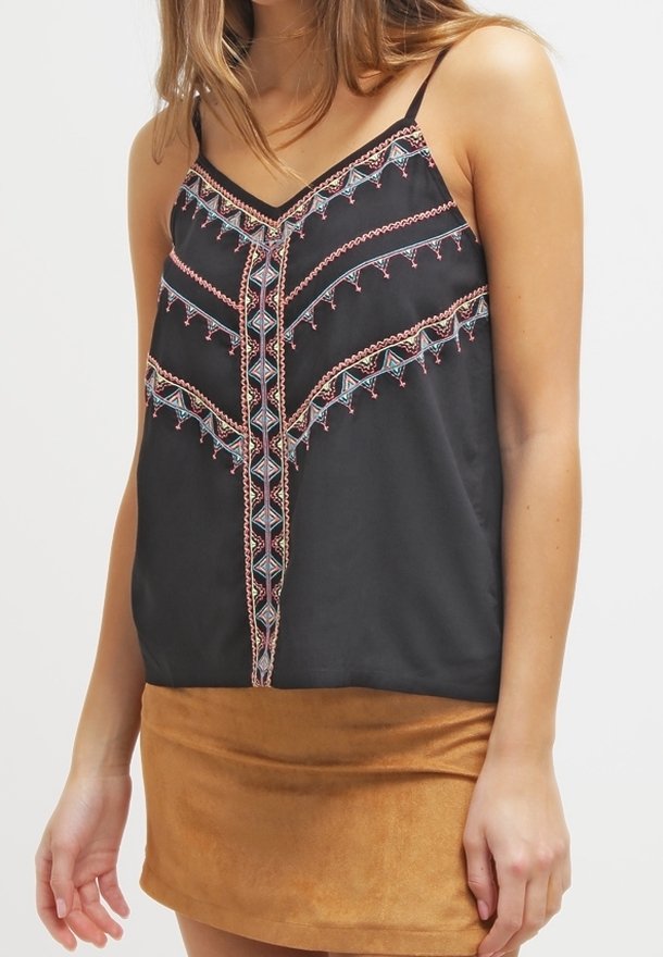 Femme portant un haut noir sans manches avec une broderie géométrique multicolore et une mini-jupe en daim beige.