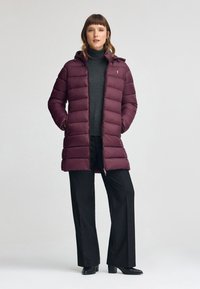 Burgunderröd puffjacka med huva, med horisontellt quiltat mönster, ståkrage och dragkedjestängning. Kombinerad med svarta byxor och klackskor.