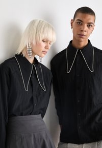 Duas modelos vestem camisas pretas com grandes colarinhos adornados com strass. Uma das modelos tem cabelo loiro comprido e brincos distintos. O fundo é neutro.