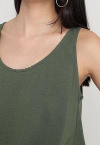 Woman wearing olive green sleeveless top with scoop neckline and large silver hoop earring, shown from neck to mid-torso.