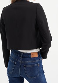 Black cropped blazer with tailored shoulders, paired with dark blue jeans featuring a brown leather tag. White shirt visible underneath.