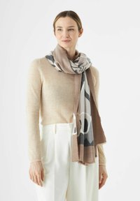 Vrouw in een beige lange mouw top, witte hoge taille broeken en een bruin-groen gestreepte sjaal om haar nek, staand voor een witte achtergrond.