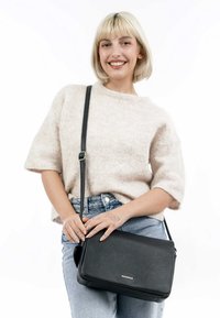 Black leather crossbody bag with a textured finish, adjustable strap, and silver hardware, worn with a light beige sweater and denim jeans.