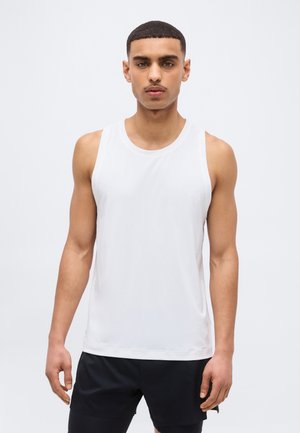 Νεαρός άνδρας με κοντά μαλλιά φοράει λευκό αθλητικό φανελάκι χωρίς μανίκια και μαύρο σορτς, στέκεται μπροστά σε απλό ανοιχτόχρωμο φόντο.