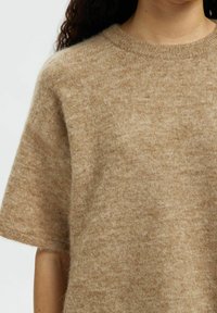 Pull en laine beige à manches courtes, col ras du cou, avec une texture douce et moelleuse. Une légère variation de lumière dans le tissu crée un look chaleureux et quotidien.