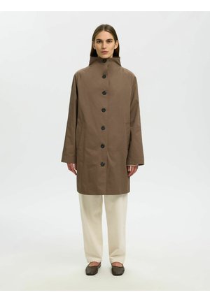 Frau steht und trägt einen braunen, geknöpften Mantel mit hohem Kragen, cremefarbene Hosen mit weitem Bein und dunkelbraune flache Schuhe vor weißem Hintergrund.