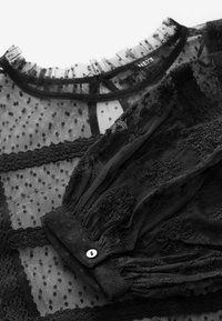 Sort gennemsigtig bluse med flæset halsudskæring, struktureret prikmønster, broderede detaljer og knapmanchetter.