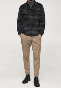 Chemise en flanelle à carreaux noirs et gris avec deux poches poitrine, associée à un pantalon beige fuselé et des bottines en cuir noir.