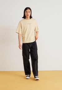 Beige oversized t-skjorte med trykk, svarte løstsittende cargo-bukser og svart-hvite sneakers på et gult gulv mot en hvit vegg.
