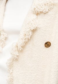 Ύφασμα με υφή κρέμας, με φουντωτές άκρες και ένα μικρό στρογγυλό μπρούντζινο κουμπί με ανάγλυφα αρχικά.