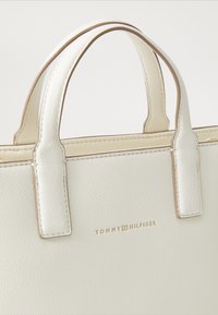 Valkoinen tekotikkukosrahkitasku, jossa on kaksoiskahvat ja kultainen Tommy Hilfiger -logo keskellä etupuolella.