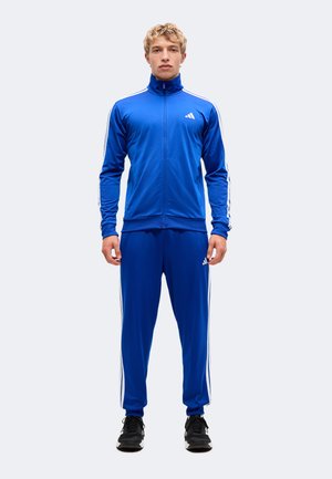Junger Mann, der einen blauen Trainingsanzug mit weißen Streifen an Ärmeln und Hosen sowie schwarze Sportschuhe trägt und nach vorne schaut.