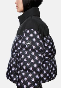 Puffjacke mit schwarzem Oberteil und einem unteren Bereich im Sternmuster in Weiß-, Rosa- und Blautönen sowie einem runden Logo-Patch am Ärmel.