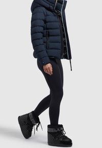 Navy donzen jack met horizontale doorgestikte patronen en een grote capuchon, gecombineerd met zwarte legging en zwarte waterdichte laarzen met voering van bont.