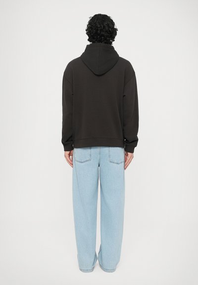 Μαύρο φούτερ με κουκούλα και άνετη εφαρμογή, σε συνδυασμό με ανοιχτόχρωμο μπλε, φαρδύ τζιν με πίσω τσέπες και ελαφρώς ξεθωριασμένη υφή.