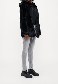 Černá bunda z umělé kožešiny s kapucí a zipem, v kombinaci se šedými slim džíny a černými kotníkovými botami na blokovém podpatku. Hladká textura, volný střih.