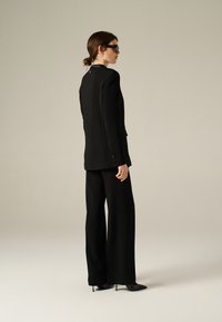 Completo nero su misura con un lungo blazer e pantaloni a gamba larga. Tessuto morbido, design strutturato, dettaglio con bottone singolo sulle maniche, tacchi alti.