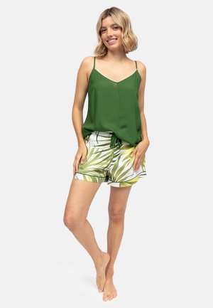 Femme souriante, portant un haut vert sans manches avec un nœud à l'avant et un short blanc imprimé de feuilles de palmier vertes et jaunes, debout pieds nus.