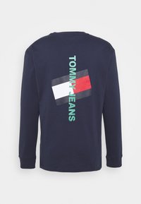 Mörkblå långärmad T-shirt med "TOMMY JEANS" tryckt vertikalt i aqua och en rektangulär logotyp i rött, vitt och svart på ryggen. Tillverkad av bomull.