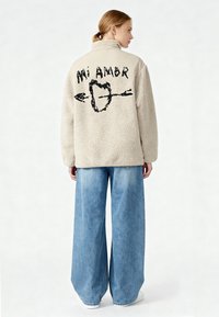Giacca in pile crema, soffice, con collo alto, caratterizzata da un motivo grafico nero sul retro che include la scritta "Mi Amor" e una freccia.
