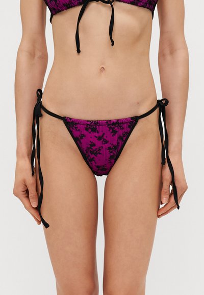 Lila bikini alsó fekete virágmintával, megkötős záródással és sima textúrával. Alacsony derékú szabású, minimális takarással rendelkezik.